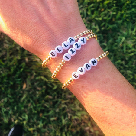Two bracelets displayed on a wrist against a grassy background, one with the name 'ELIZABETH' and the other with 'IAN' written in black acrylic letter beads among gold filled beads.