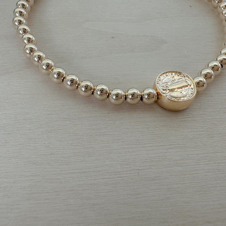 A gold-filled bracelet with St. Benedict coin bead, featuring a series of round beads strung on an elastic cord.