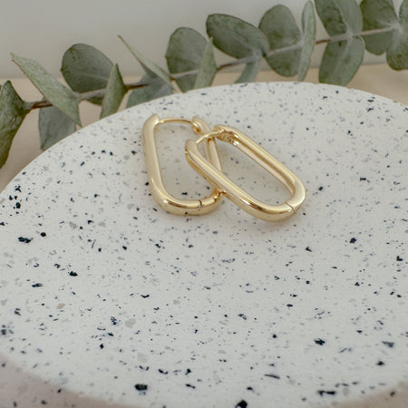 A pair of gold-filled link hoop earrings on a textured white stone with a green leaf in the background.