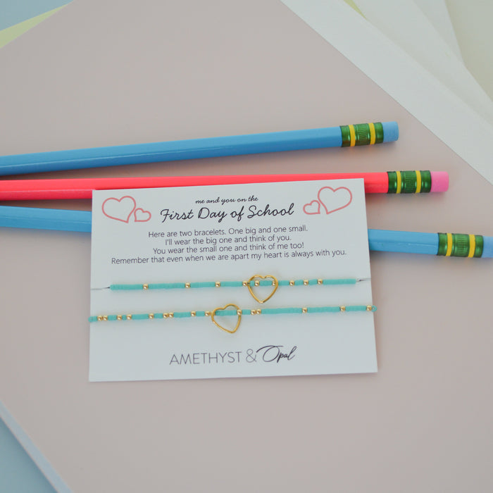 A close-up of a gift card featuring two delicate beaded bracelets, one light blue with gold beads and one gold with a heart charm, displayed on pastel-colored notebooks with blue and pink pencils.