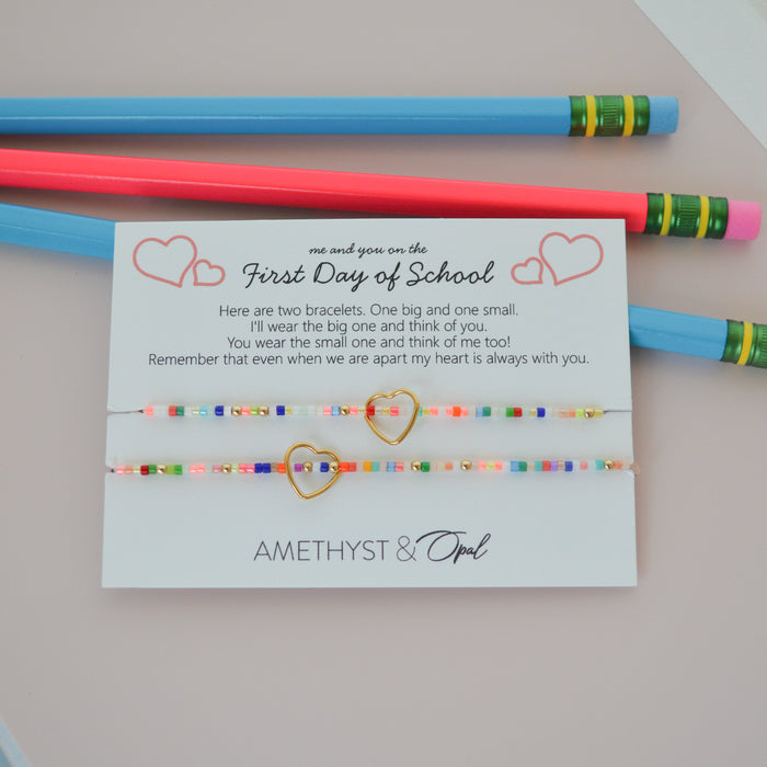 Two colorful beaded friendship bracelets with gold heart charms, presented on a white gift card with school-themed text and decorated with two pencils on a light pink background.