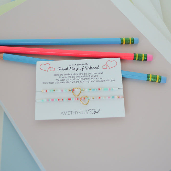 A gift set for first day of school featuring two colorful beaded friendship bracelets on a white card, styled with two pencils on pastel-colored paper sheets.