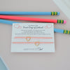 A first day of school gift set featuring a delicate gold heart-themed cord bracelet on a white card, styled with two colorful pencils (one pink, one blue) on a pastel-colored paper background. The brand 'AMETHYST & Girl' is visible on the card.