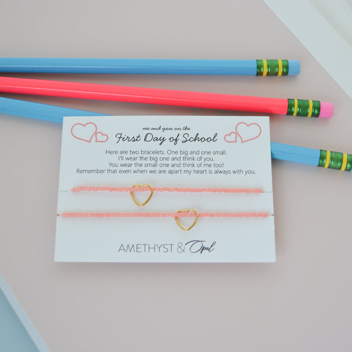 A first day of school gift set featuring a delicate gold heart-themed cord bracelet on a white card, styled with two colorful pencils (one pink, one blue) on a pastel-colored paper background. The brand 'AMETHYST & Girl' is visible on the card.