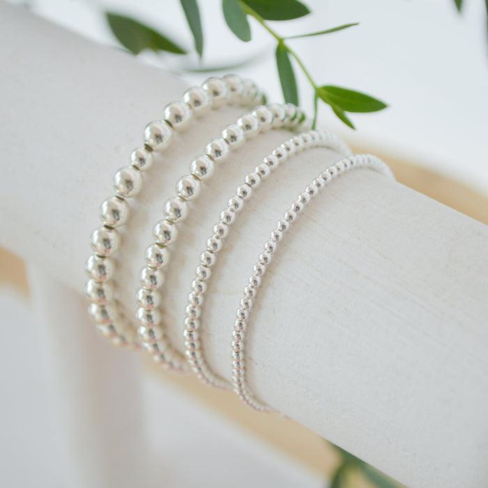 Silver beaded bracelets on a white surface with green leaves in the background