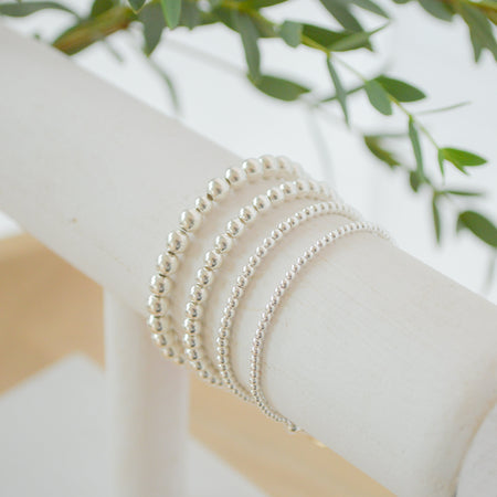 Silver beaded bracelets on a white surface with green leaves in the background