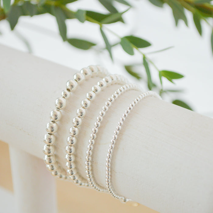 Silver beaded bracelets on a white surface with green leaves in the background